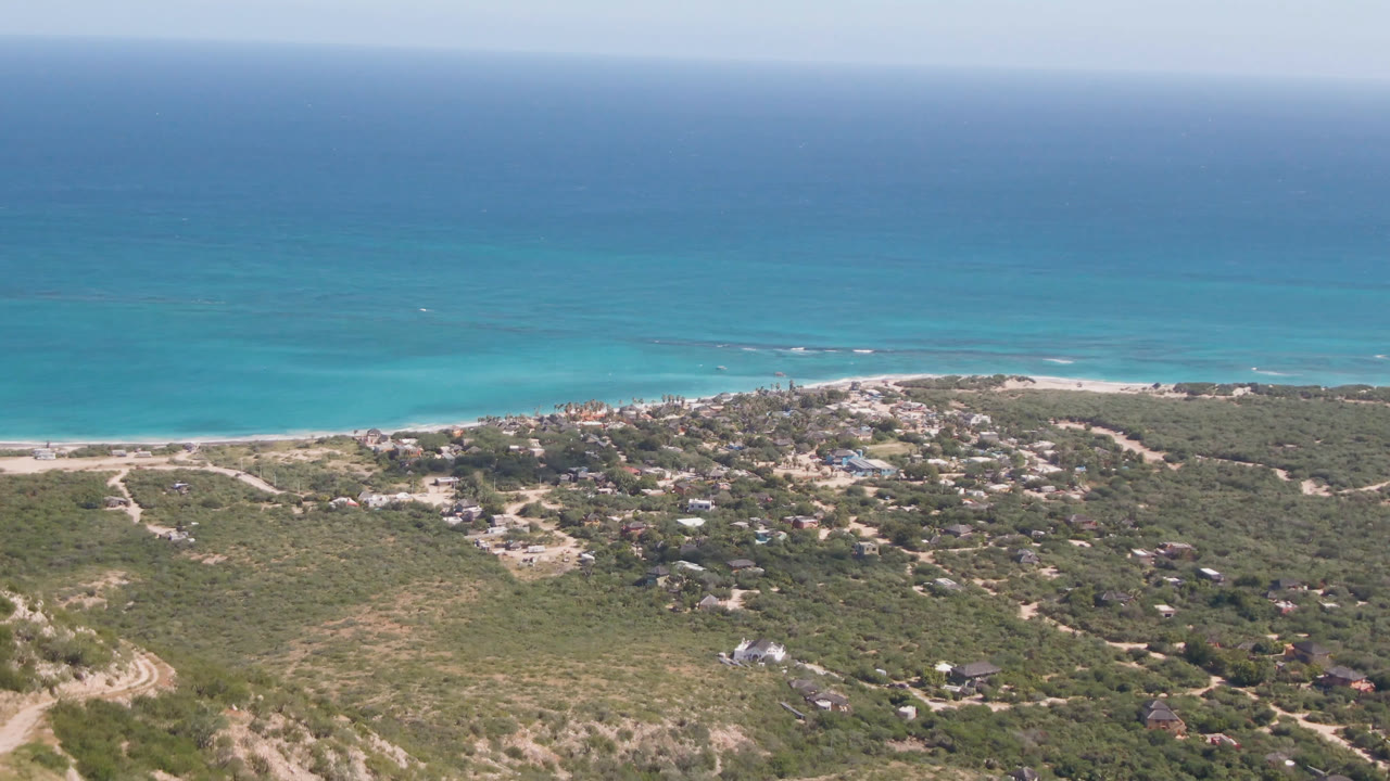Aerial coastline with blue water, sandy shore, brushy terrain, and scattered houses.