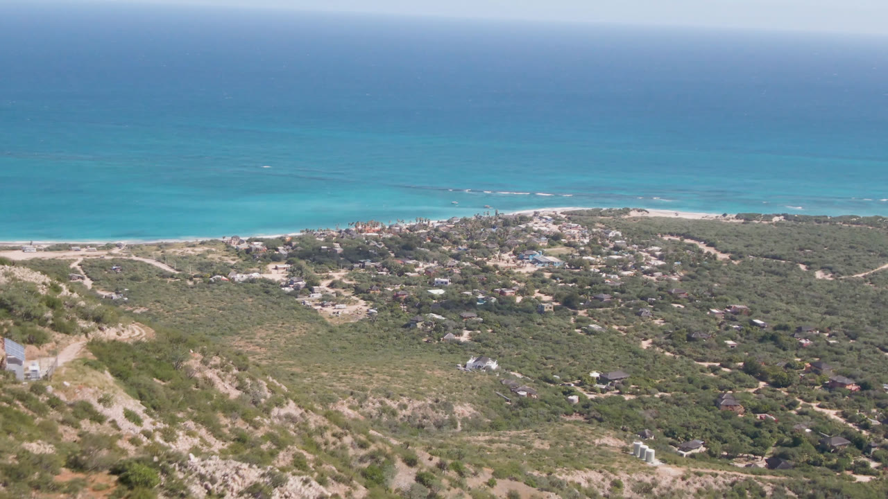 Opening frame of Cabo Pulmo coastline with turquoise water, a pale beach strip, scrub-covered hills, scattered distant homes, rocky foreground, and no visible people.