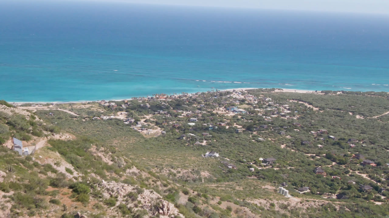 Cabo Pulmo hillside view at 0:01 with the ocean horizon high in frame, beach below it, green coastal terrain, distant houses, and no visible people.