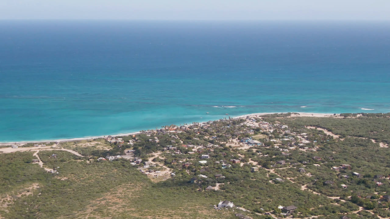 Aerial view of blue ocean, sandy beach, scrubland, and a small coastal town.