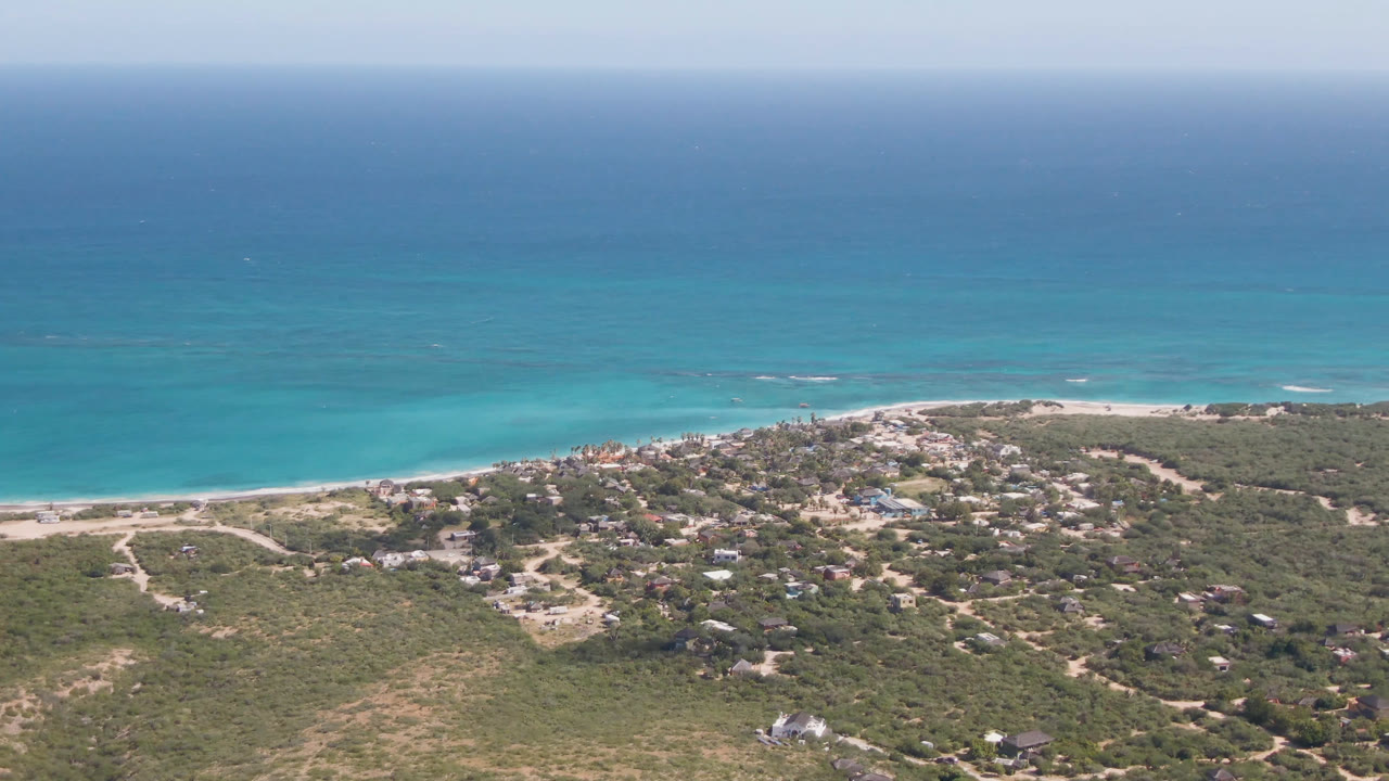 Aerial view one second into the Cabo Pulmo clip, showing the beach curving below turquoise water, clustered small buildings inland, dry vegetation, and no visible people.