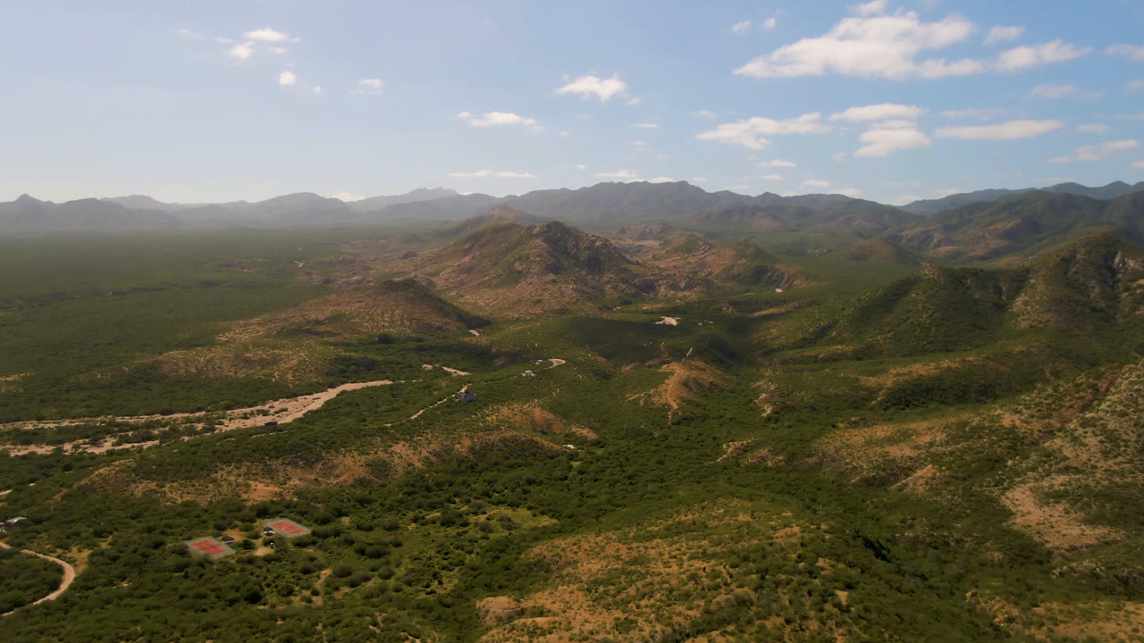 Wide aerial view of desert hills, scrub, a pale wash, and distant mountains.