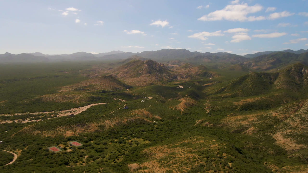 Aerial frame from Cabo Pulmo with scrub-covered foreground, a pale riverbed cutting through the valley, rugged midground hills, hazy mountains, and no visible people.