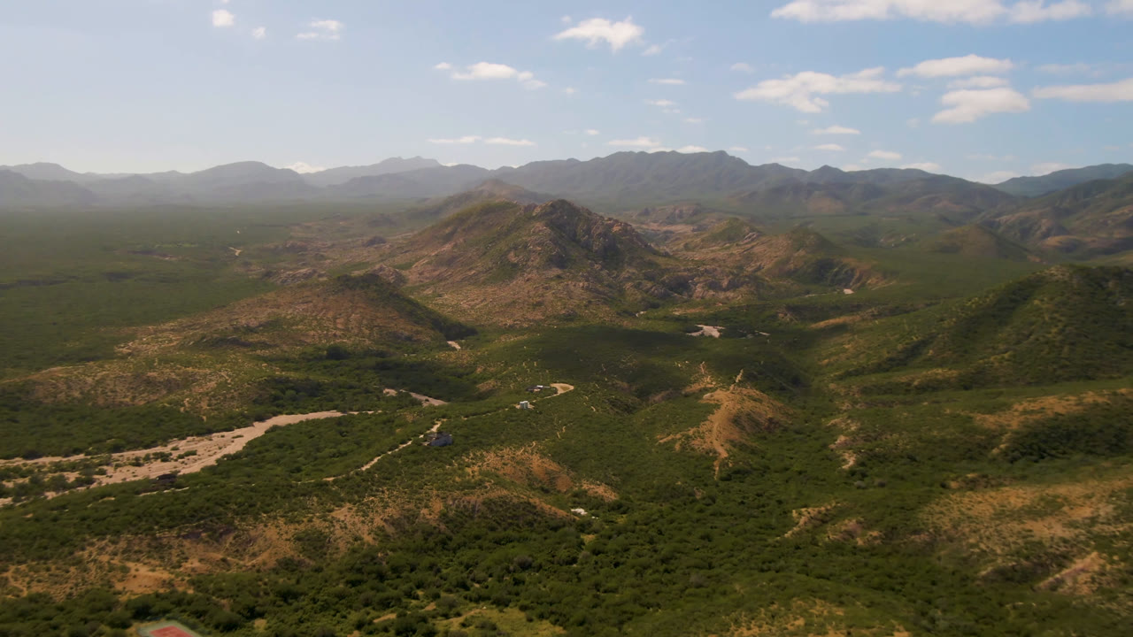 Wide aerial frame over Cabo Pulmo, Baja California Sur, showing green valley vegetation, a pale winding river wash, rocky hills, hazy distant mountains, and no visible people.