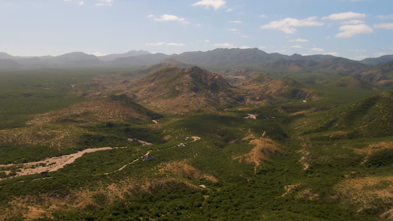 Aerial view in Cabo Pulmo with the rocky ridge slightly more centered, dense green vegetation below, a light riverbed crossing the valley, hazy background mountains, and no visible people.