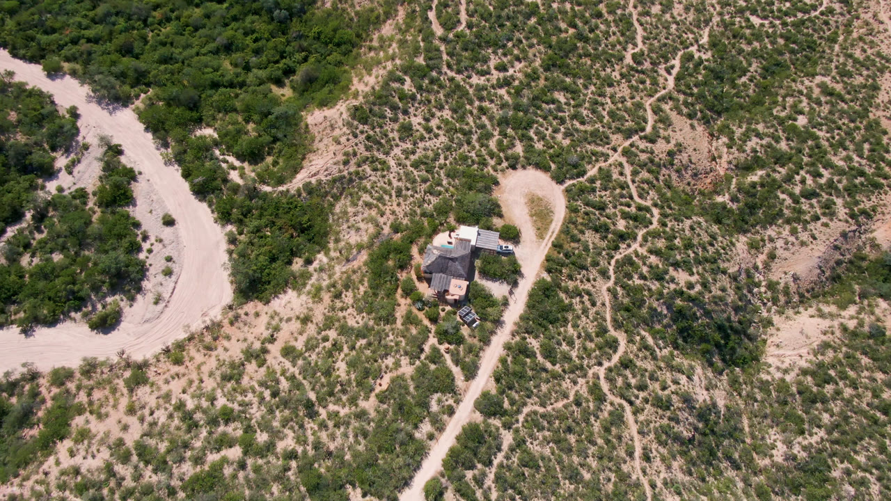 Wide drone view in Cabo Pulmo, Baja California Sur, with the house slightly left of center, a winding dirt track beside it, dense green trees, tan ground, and no visible people.