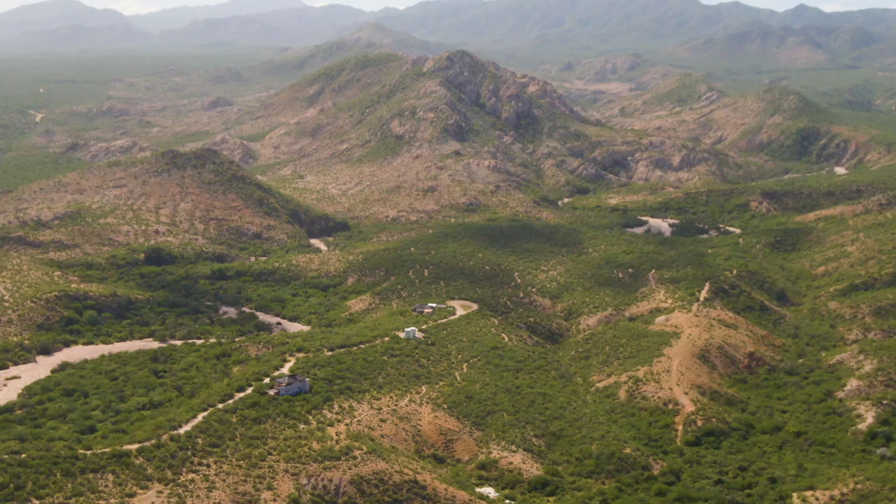 Opening aerial frame over Cabo Pulmo, Baja California Sur, Mexico, with rocky hills, dense green scrub, a winding dirt road, a pale wash, scattered small buildings, and no visible people.
