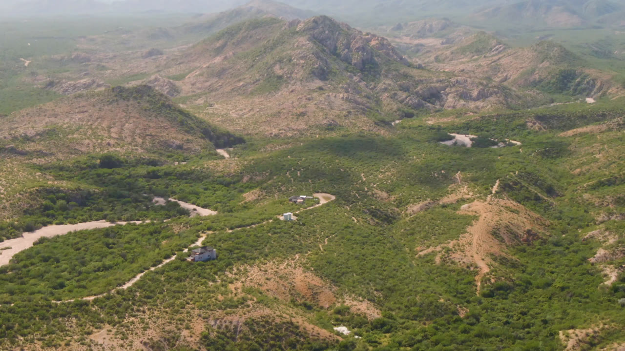 Second aerial frame in Cabo Pulmo shows the curved dirt road nearer the lower frame, green vegetation across the valley floor, rocky hills behind it, and no visible people.