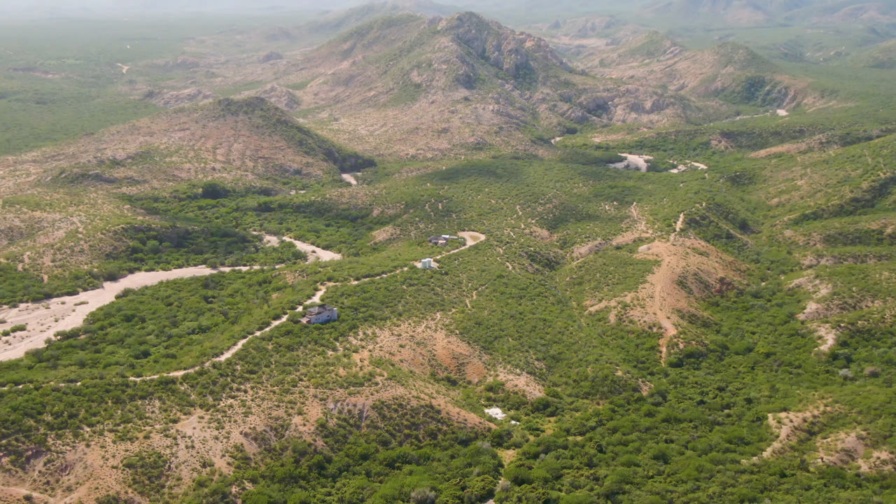 Wide aerial frame from Cabo Pulmo, Baja California Sur, showing scrub-covered hills, a pale winding riverbed, a narrow dirt road, distant ridges, and no visible people.