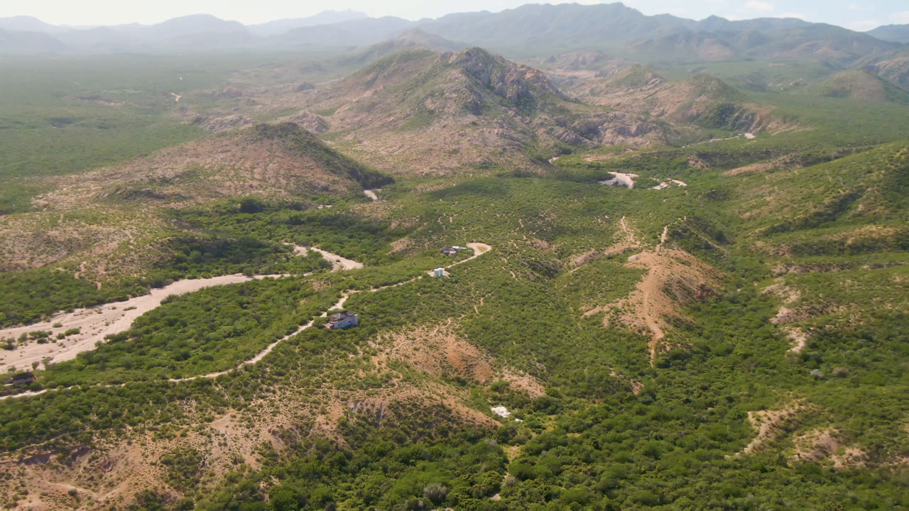 Drone view across a green valley, rocky mountain slopes, a dirt road, and a pale wash.