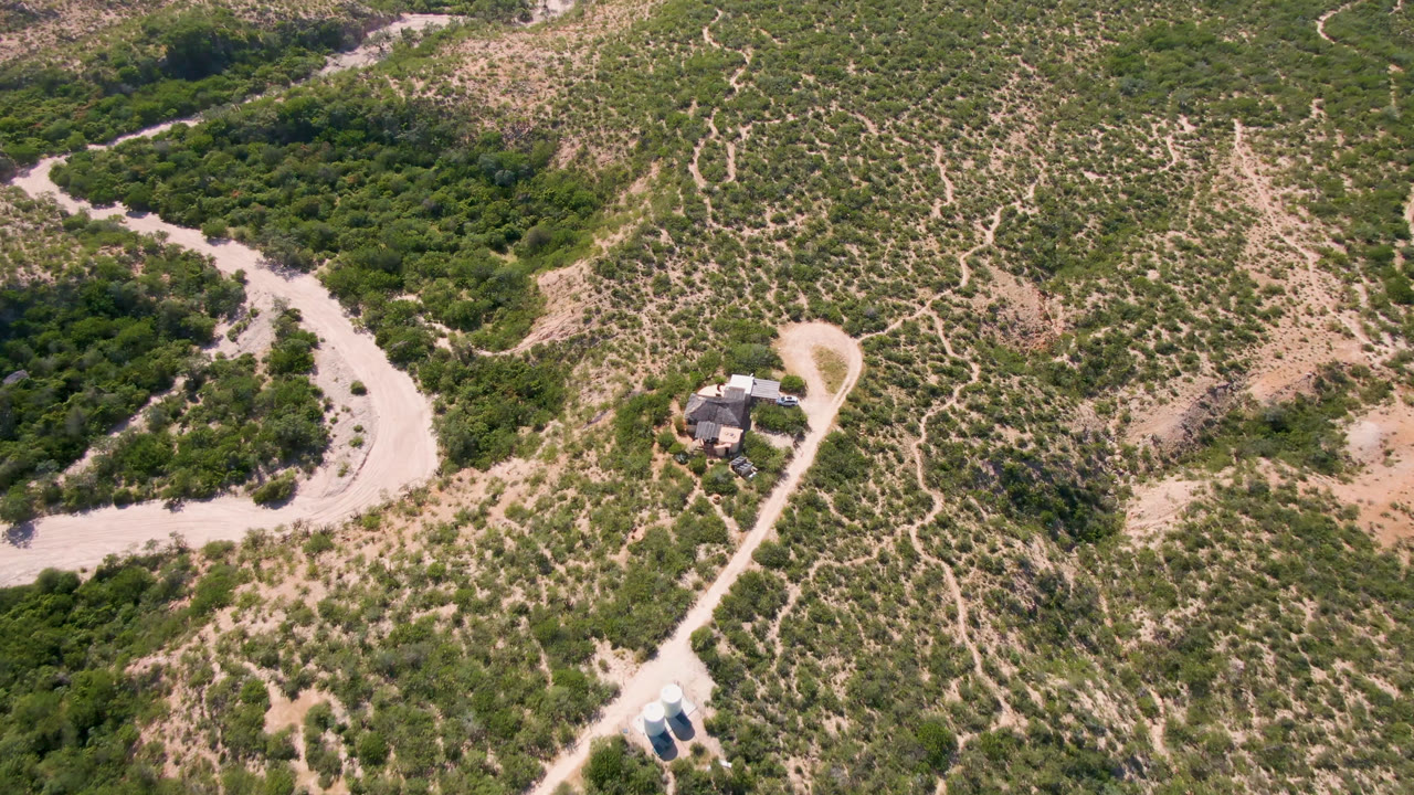 Second aerial frame in Cabo Pulmo holds the house slightly lower in view, with the looping access track, dense green vegetation to the left, dry footpaths, and no visible people.