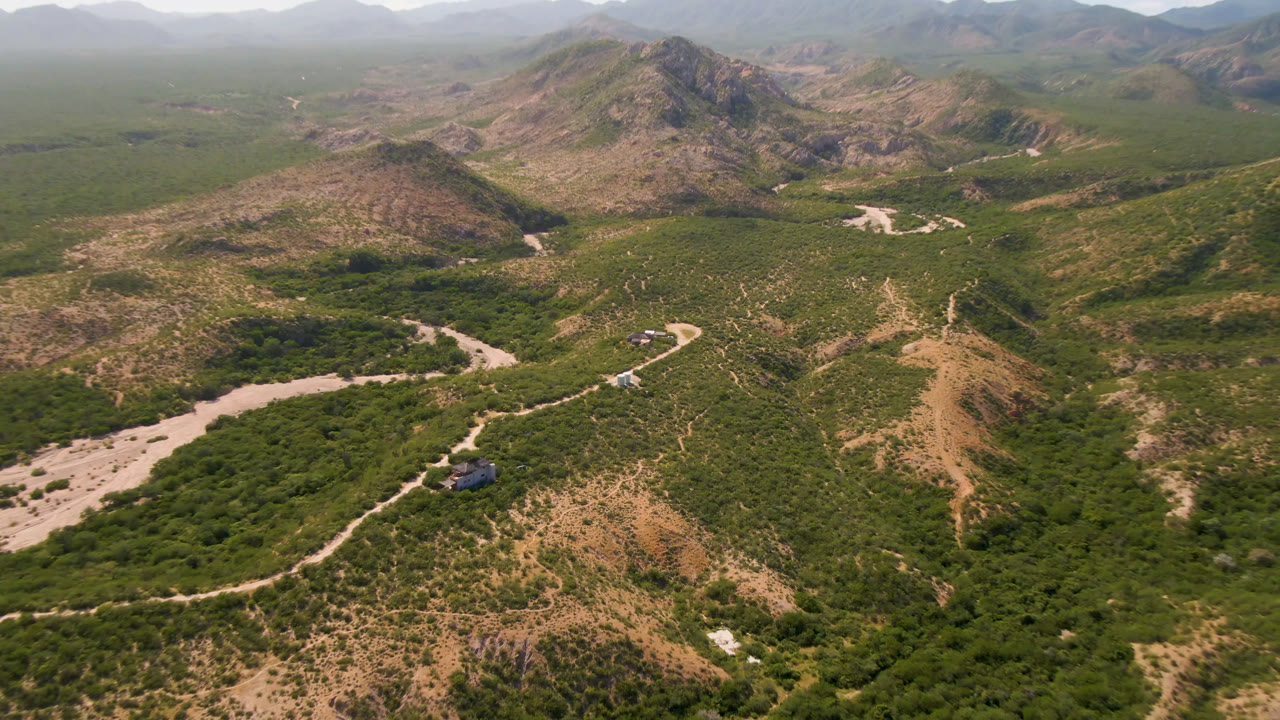 Aerial frame near Cabo Pulmo showing the dry wash more centrally, with the winding road crossing green vegetation, tan rocky slopes, distant mountains, and no visible people.
