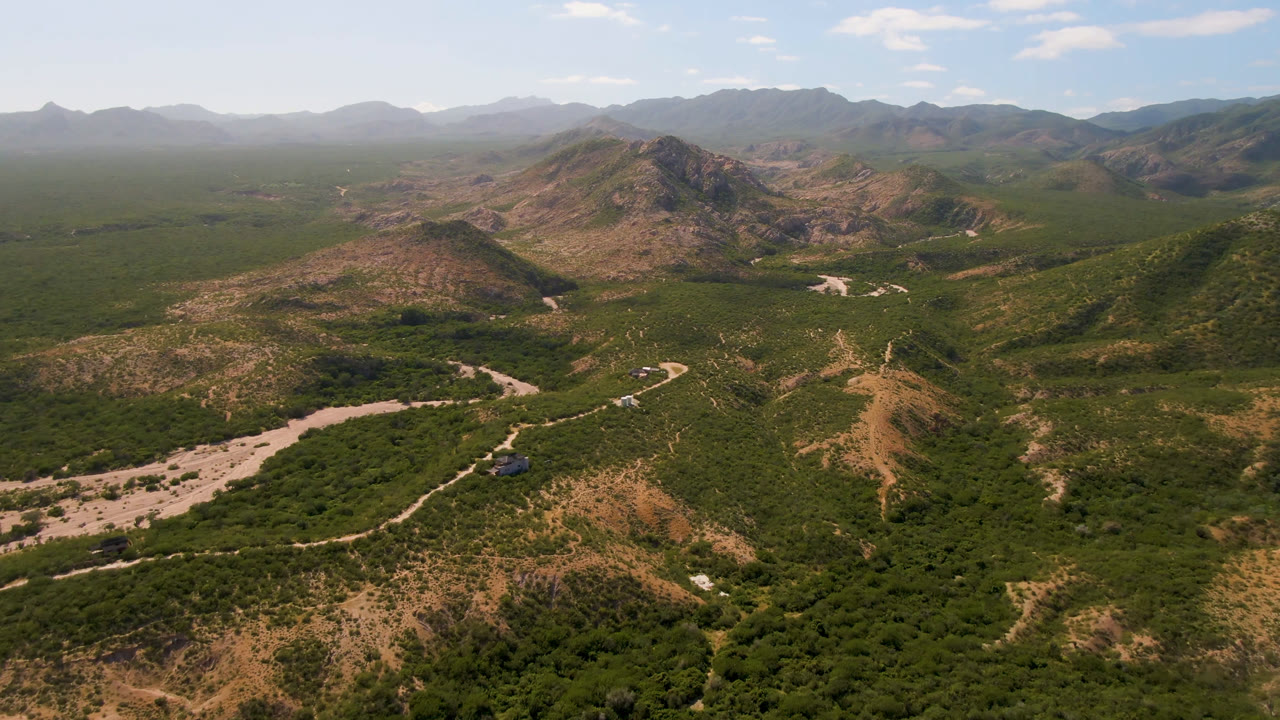 A wide Cabo Pulmo aerial shows a pale dirt road and dry wash crossing green brush below rocky ridges, with hazy mountains in the distance and no visible people.