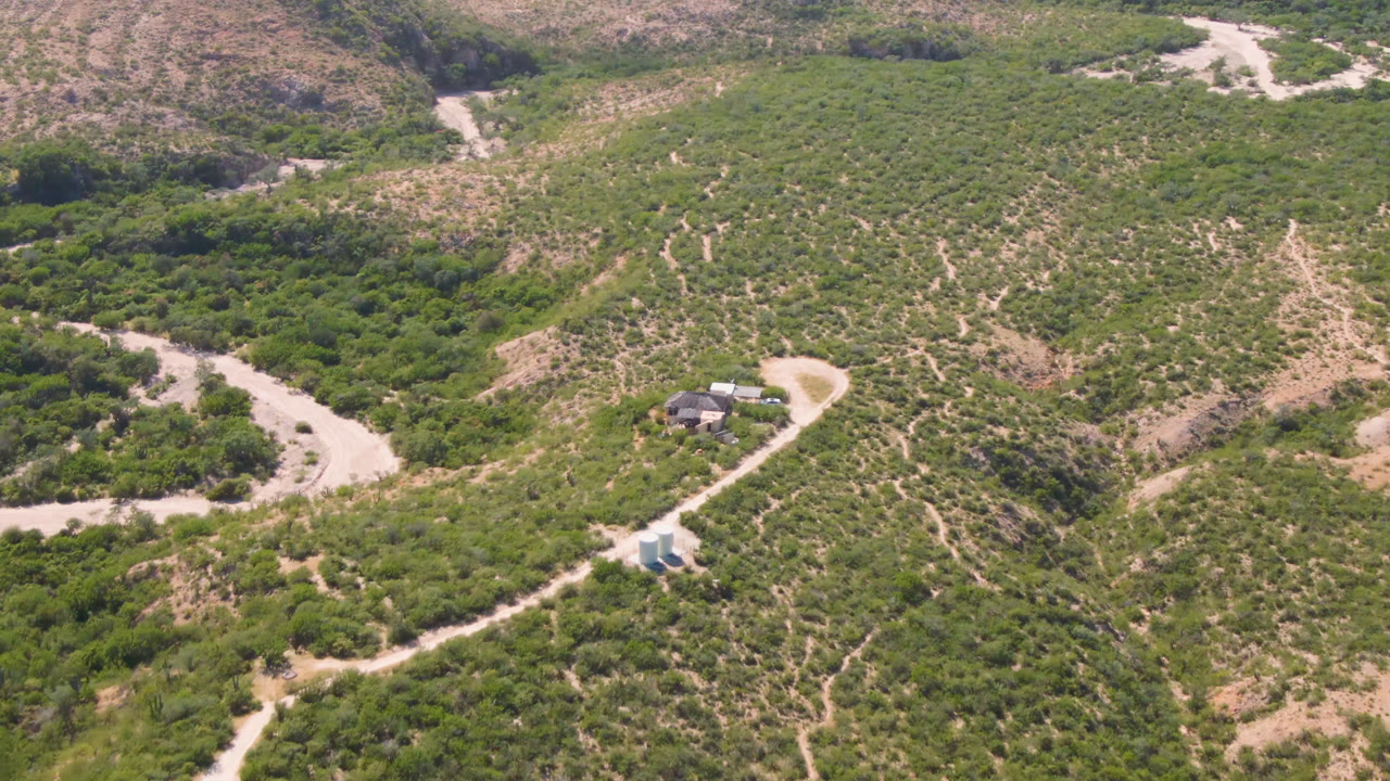 Opening aerial frame over Cabo Pulmo terrain shows a small house near a pale dirt road, dry wash bends, green scrub, tan hillsides, and no visible people.