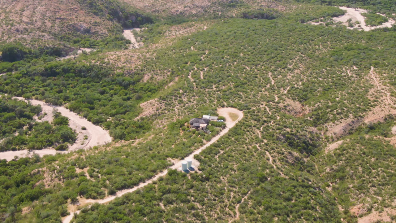 At 0:01, the Cabo Pulmo aerial view holds the small house near the lower center, with a winding dirt road, dry channel, scrubby slopes, and no visible people.