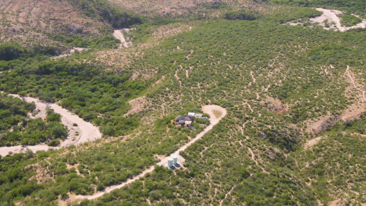 Wide aerial view of a hillside house, pale dirt road, and green scrub.