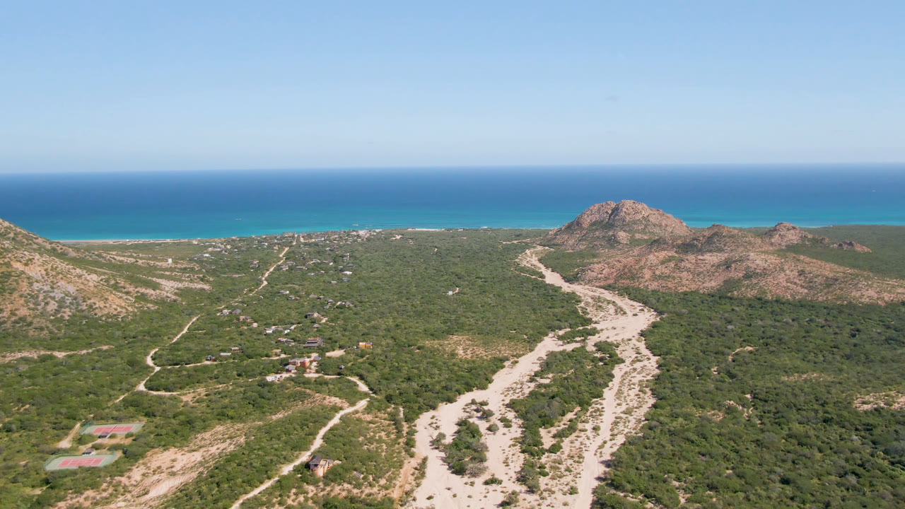 Cabo Pulmo aerial view with a winding pale road through green scrub, rocky hills to the right, turquoise shoreline, and no visible people.