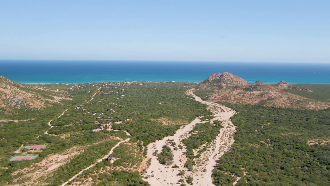 Aerial view of winding dirt roads through green scrub toward rocky hills and ocean