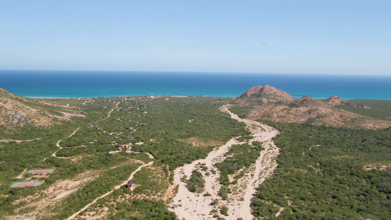 Aerial frame in Cabo Pulmo with branching dirt tracks and small structures in the desert foreground, rocky hills to the right, ocean beyond, and no visible people.