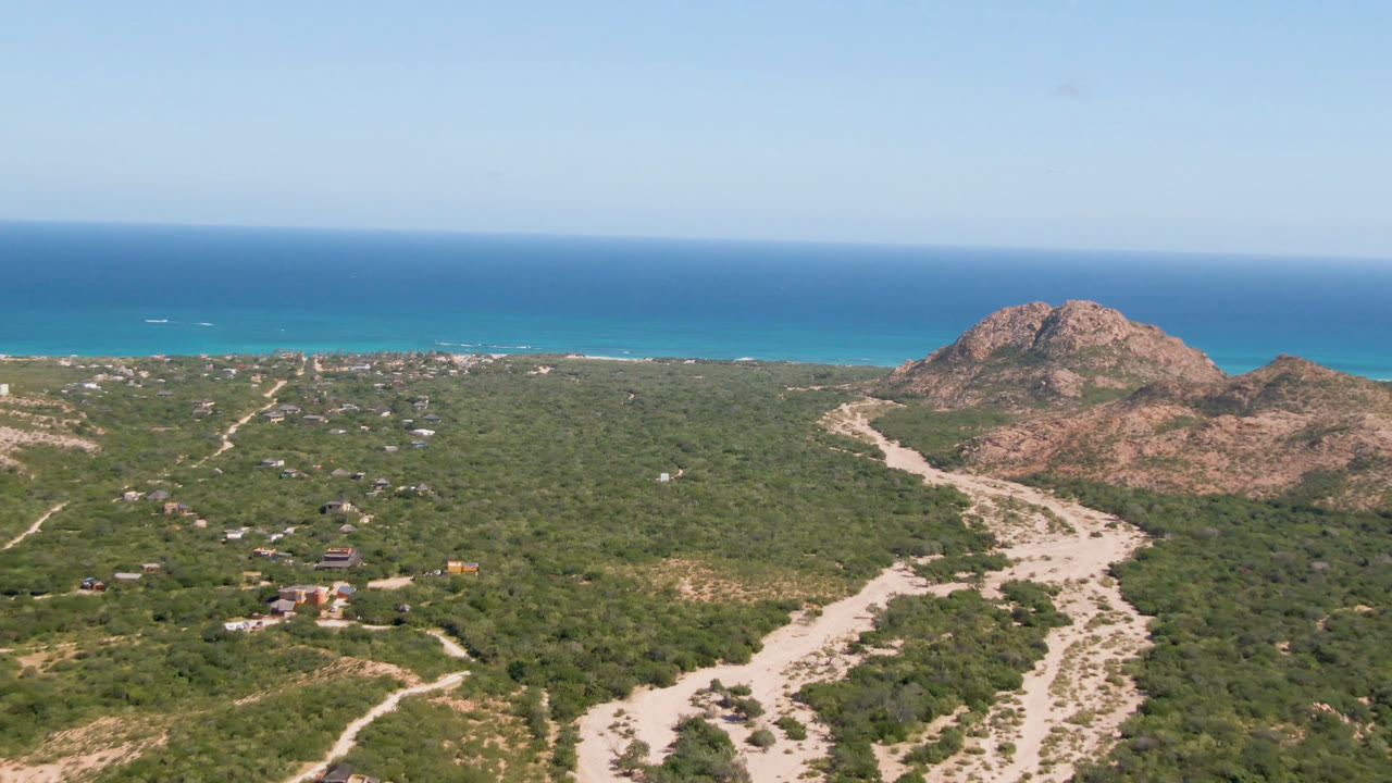 Aerial view over Cabo Pulmo showing green desert scrub, pale sandy washes, reddish rocky hills, blue ocean, clear sky, and no visible people at the start of the clip.