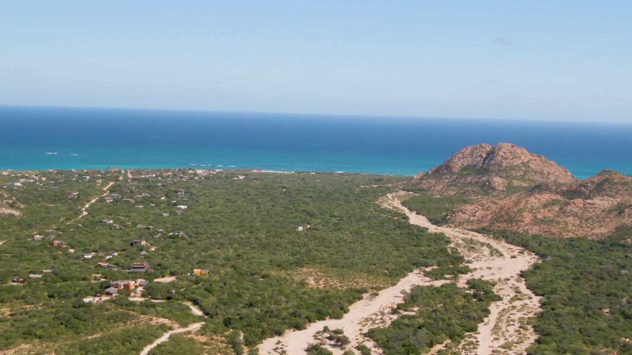 Wide aerial frame from Cabo Pulmo with a central rocky hill, dry wash channels through scrubland, scattered small structures, blue ocean, and no visible people.