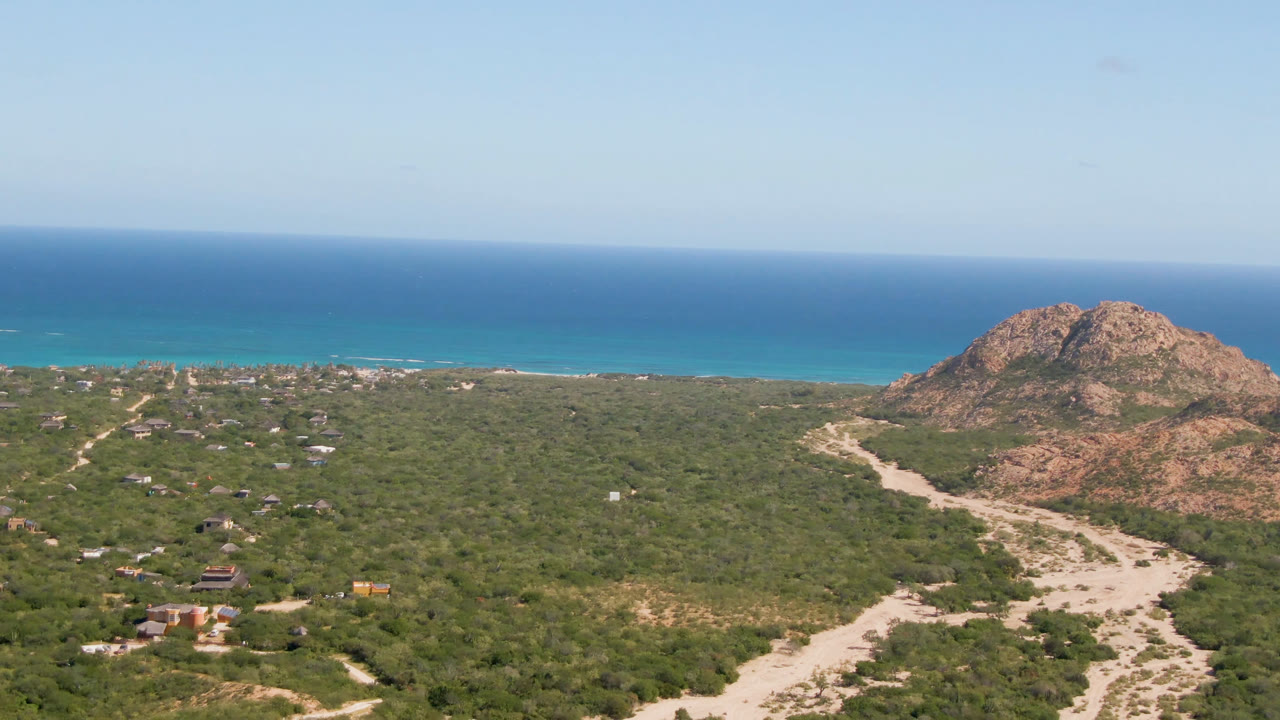 Opening aerial frame over Cabo Pulmo scrubland with a pale dirt road leading toward rocky hills, turquoise shoreline, blue ocean, clear sky, and no visible people.