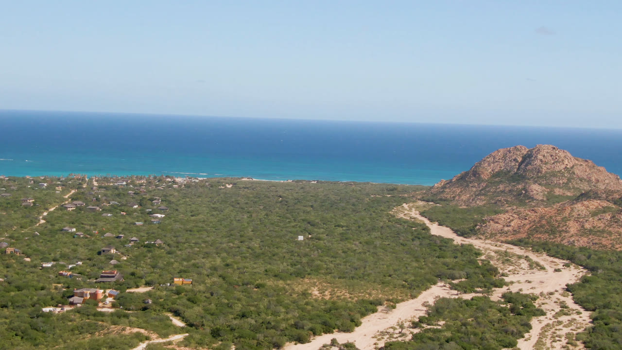 Aerial view in Cabo Pulmo with the winding sandy track more centered through green scrub, rocky hills to the right, ocean across the background, and no visible people.