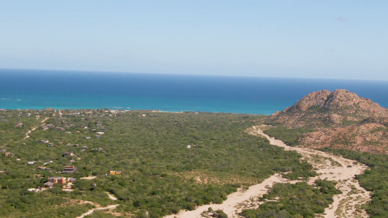 Drone view of desert vegetation, pale road, rocky hill, turquoise coast, and open sea.