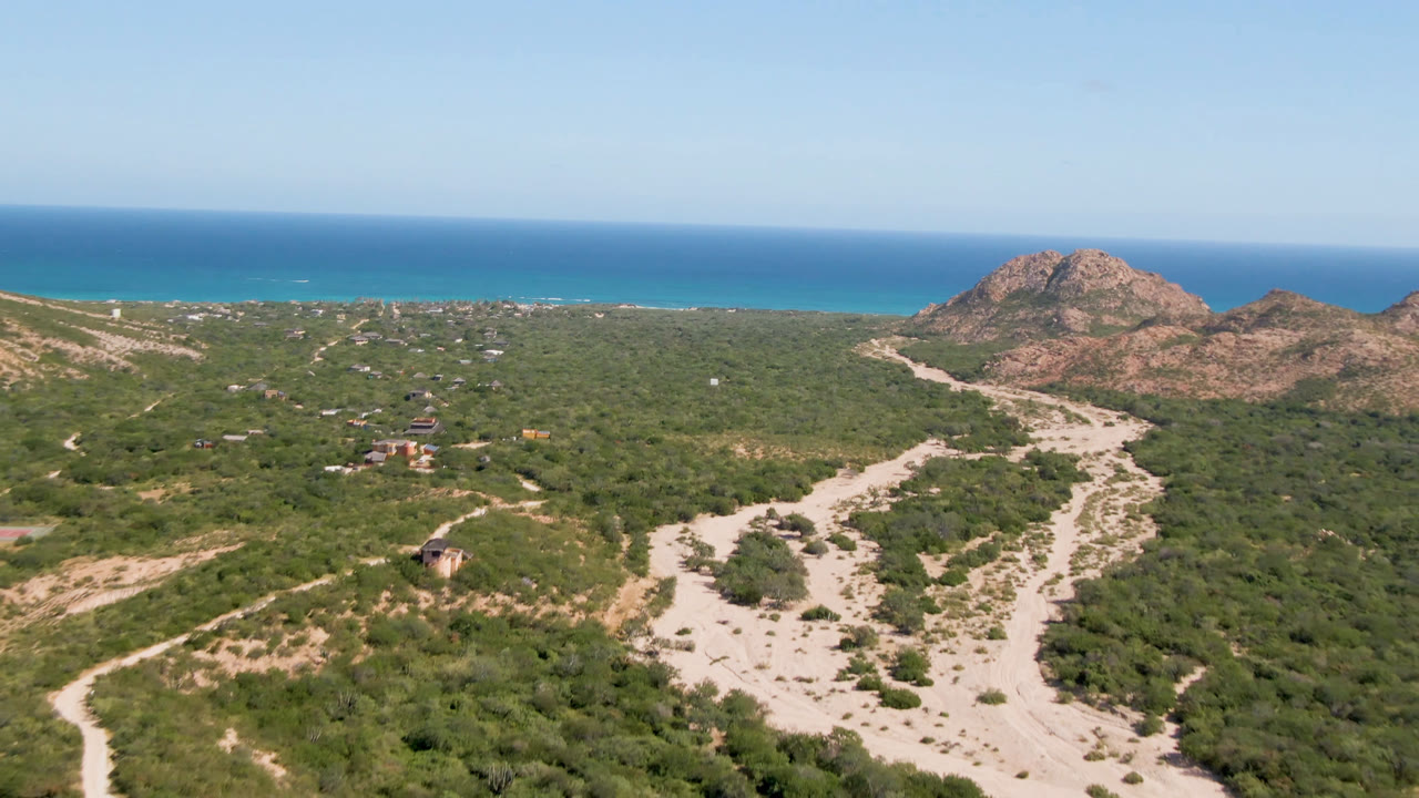 Aerial view in Cabo Pulmo with a broad pale wash or road crossing the foreground, small structures in the scrub, rocky hills at right, blue sea behind them, and no visible people.