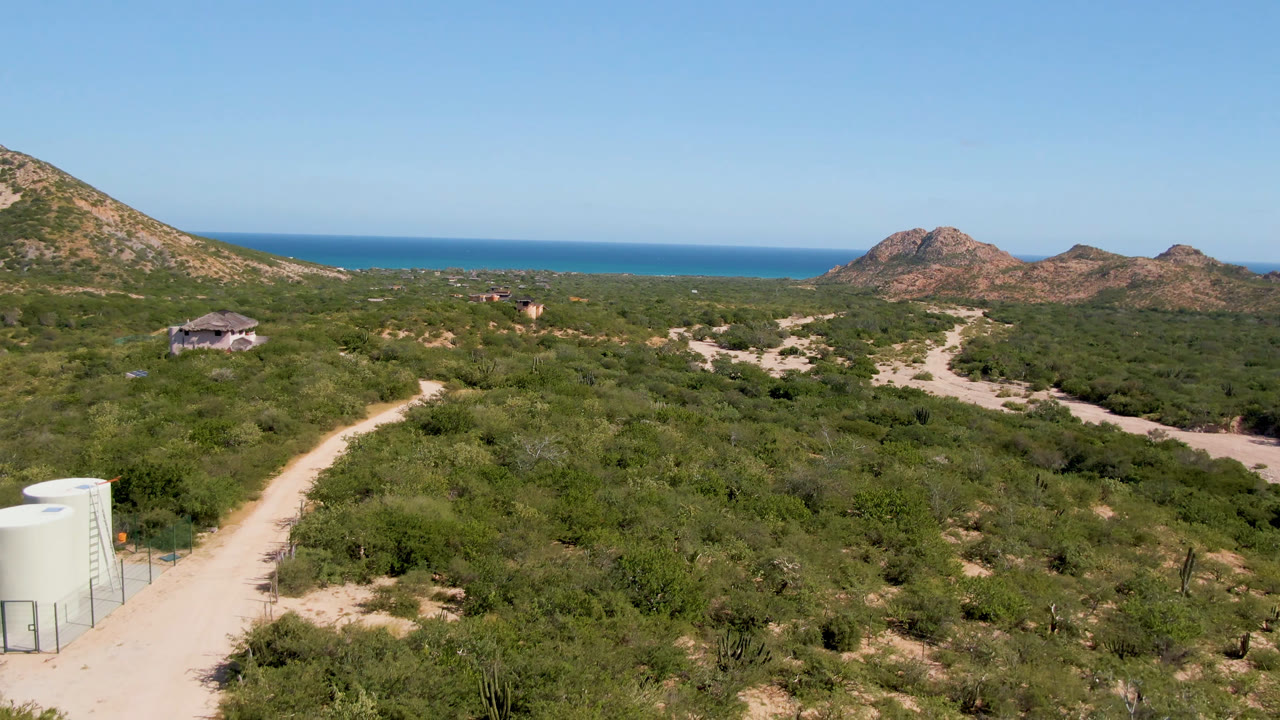 Cabo Pulmo frame with the dirt road more centered, white cylindrical tanks at the lower right, scrubland across the midground, rocky hills, ocean, and no visible people.