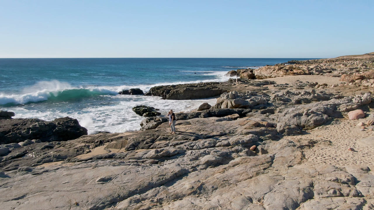 The two walkers are slightly farther along the rocks as a wave curls and breaks across the midground shoreline at Laguna Beach near San Jose Del Cabo, Mexico.