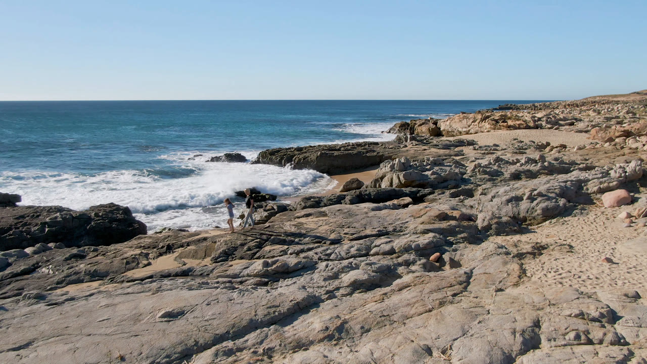 Wide view of Laguna Beach in San Jose Del Cabo, Mexico, with two people on the left side of the rock shelf as waves break against dark coastal rocks.