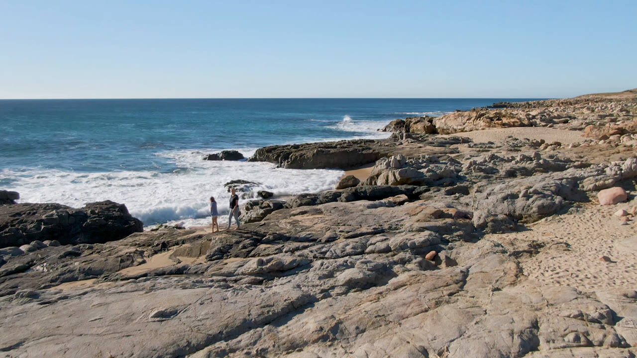 At 0:01, two people stand closer to the center-left rocks at Laguna Beach in San Jose Del Cabo, Mexico, with white surf spreading behind them.