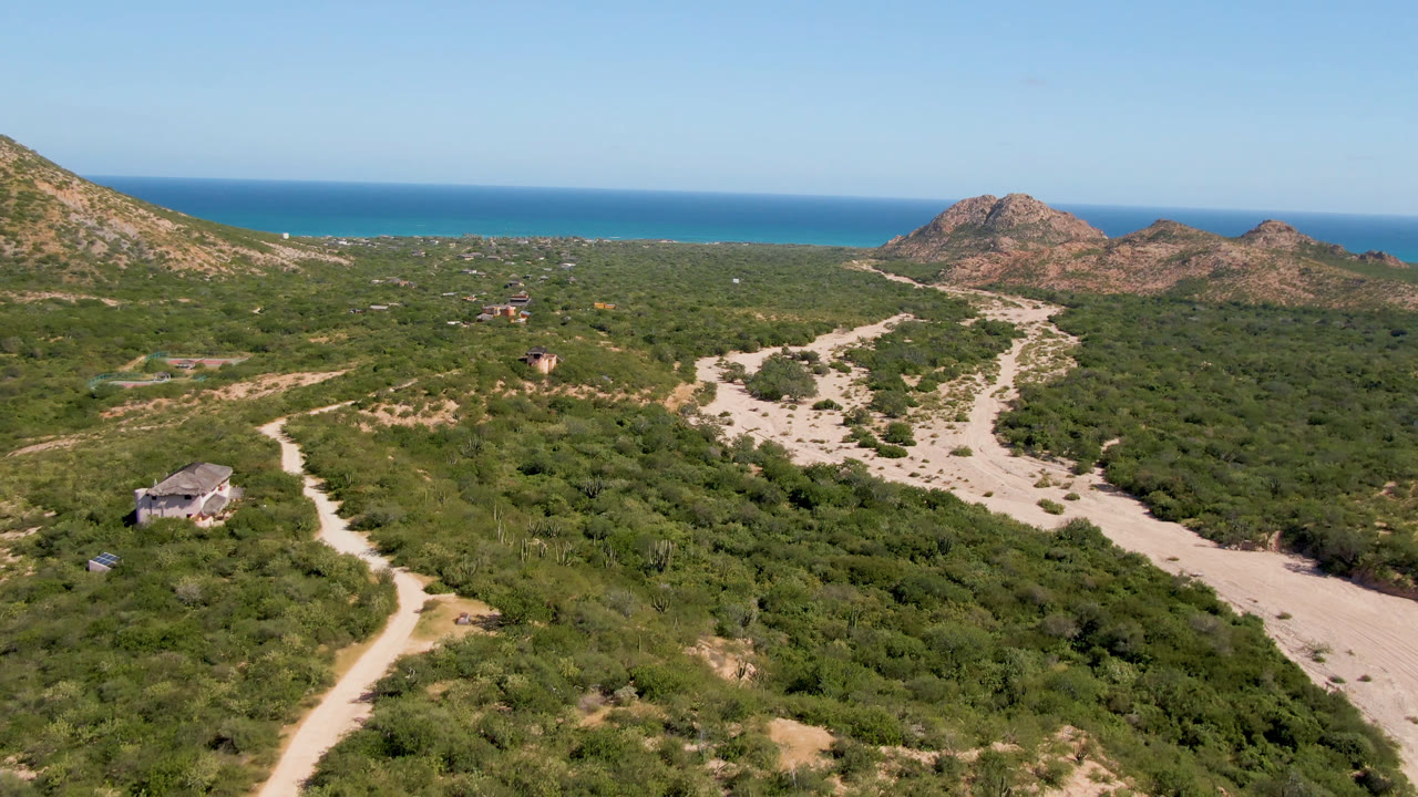 Aerial view of a dirt road crossing green scrubland toward rocky hills and ocean.