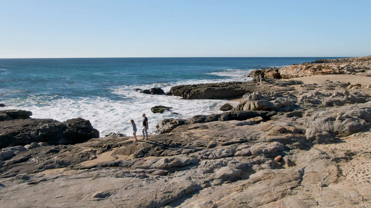 Two people walk across pale rocks at Laguna Beach in Los Cabos, Mexico, with white surf rolling in behind them and a clear blue sky above.