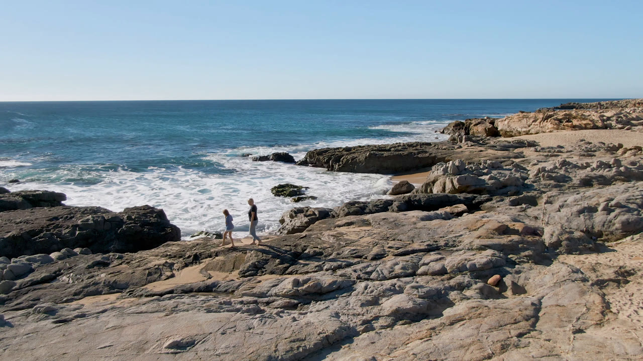 The walkers have moved slightly right across the Baja rocks while foamy water spreads around dark outcrops near the shore.