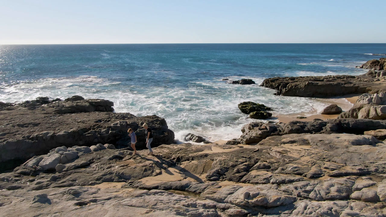 Two people stand on the rocky shoreline at San Jose Del Cabo, Mexico, with white surf breaking behind them and long shadows stretching across the foreground rocks.