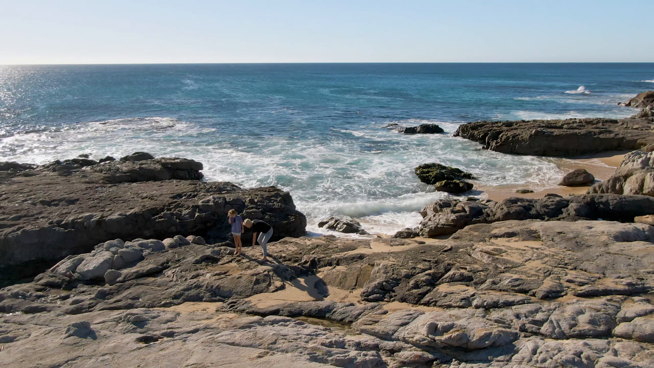 An adult and child pause on pale shoreline rocks in San Jose Del Cabo, Mexico, while foamy waves roll through the rocky coves behind them.