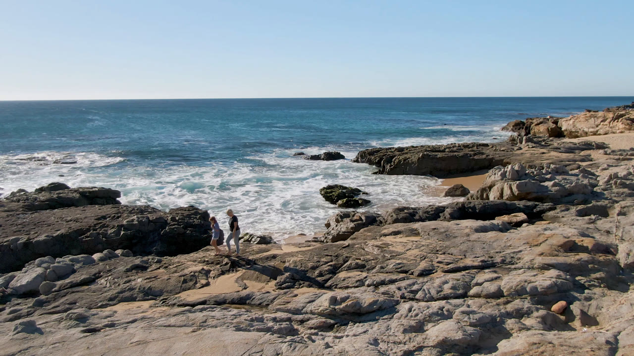 Two people walk across pale rock shelves on the San Jose Del Cabo coast as white surf breaks behind them and the blue horizon spans the frame.