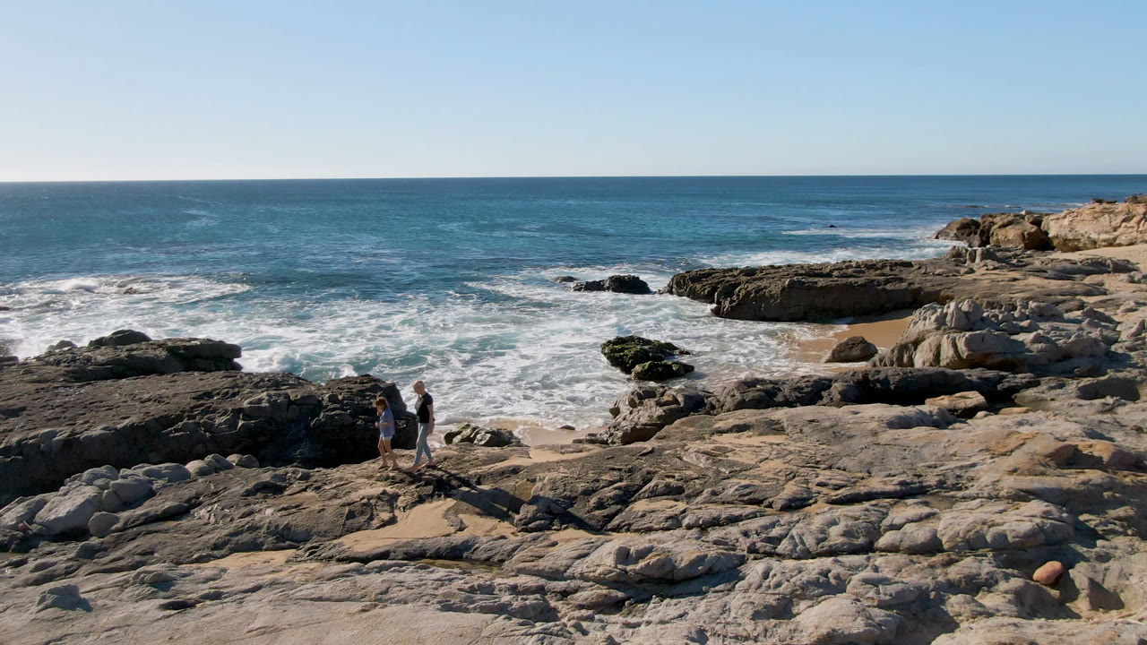 The walkers move along the rock ledge near tide pools in San Jose Del Cabo, with foamy waves spreading across the midground and offshore rocks beyond.