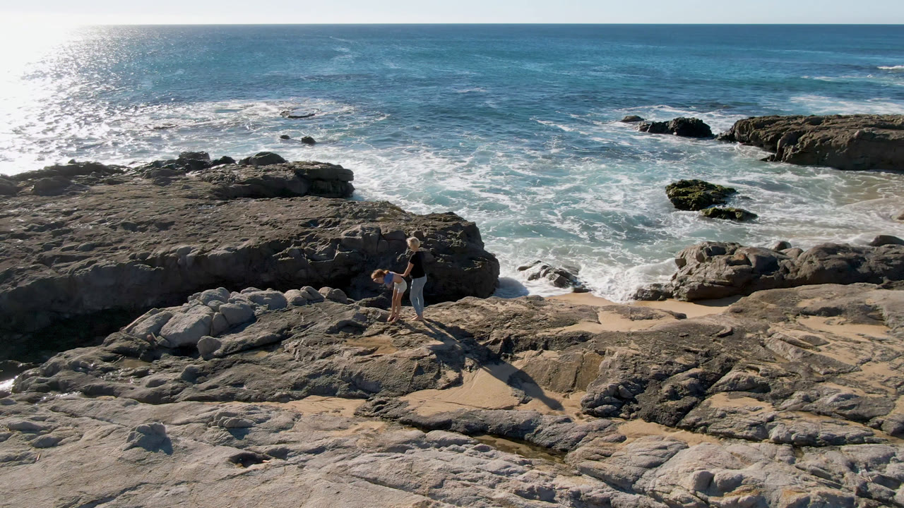 At San Jose Del Cabo, Mexico, the two figures remain on the midground rocks while white water spreads across the beach and dark rocks sit offshore.