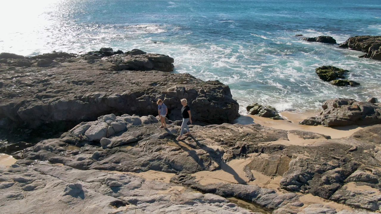 Two people walk across pale rocks at Laguna Beach in San Jose Del Cabo, Mexico, with bright ocean glare, white surf, and dark offshore rocks behind them.