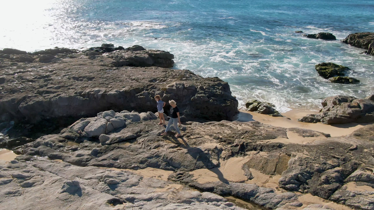 Two people stand farther along the rock shelf in San Jose Del Cabo, Mexico, while foam curls around sand channels and low dark rocks in the water.