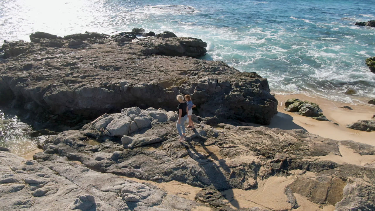 Two people walk across pale rock at Laguna Beach in Los Cabos, Mexico, with white surf breaking behind a dark rock shelf and long shadows stretching across the foreground.