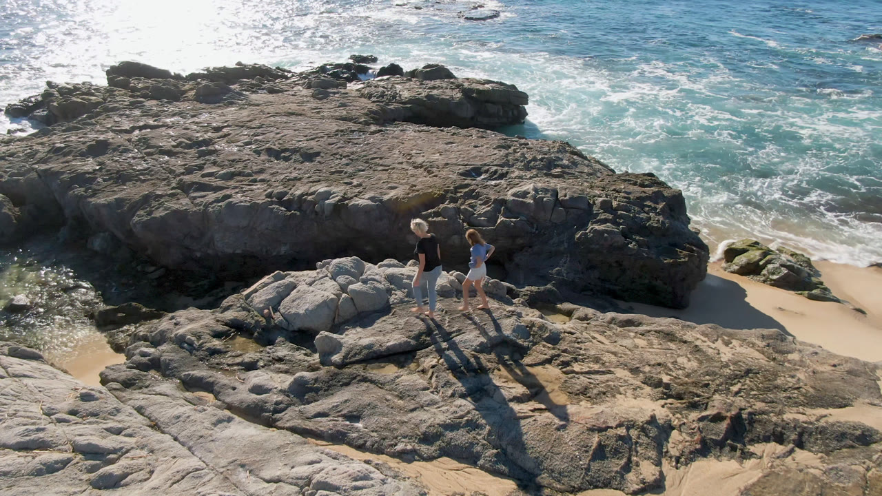 Two people stand or step across uneven coastal rock at Laguna Beach in Los Cabos, Mexico, with dark stone ledges, pale sand, and churning blue-green surf behind them.