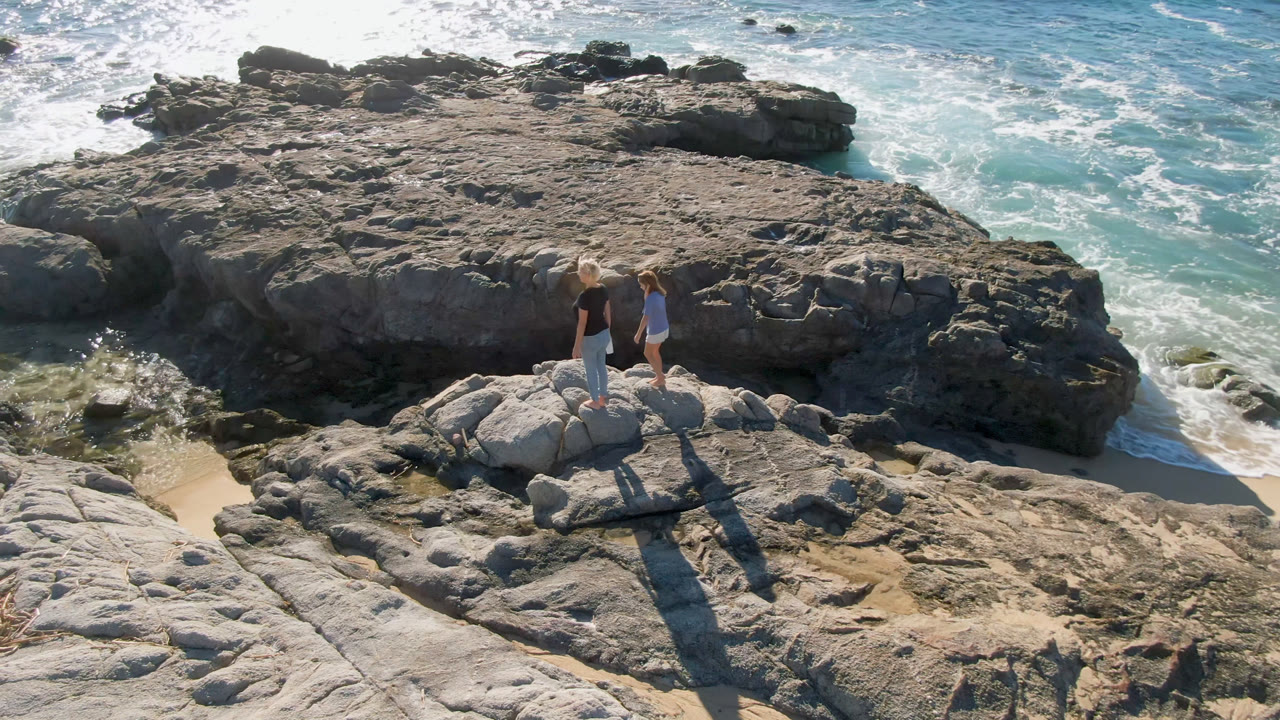 At Laguna Beach in Los Cabos, a woman and child stand on pale rocks while foamy turquoise water moves around darker coastal outcrops.