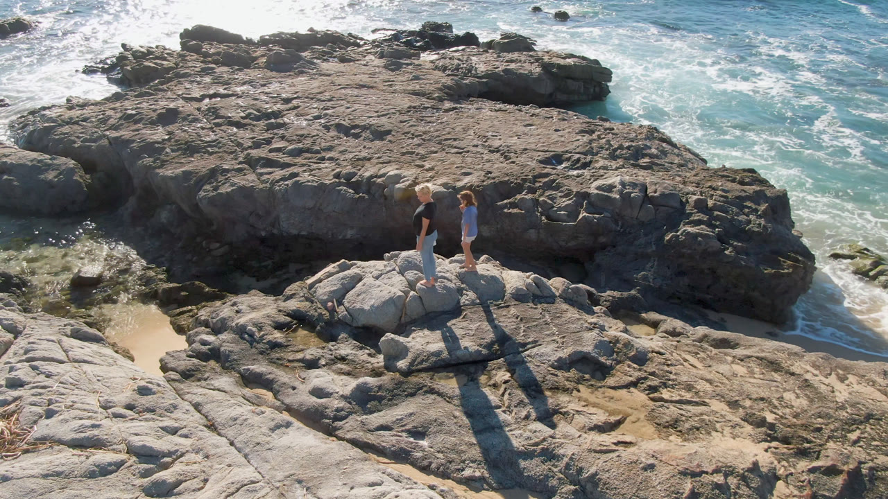 At 0:01, the woman and child remain on the rock ledge as white water curls through tide pools beside the darker shoreline rocks in Los Cabos.
