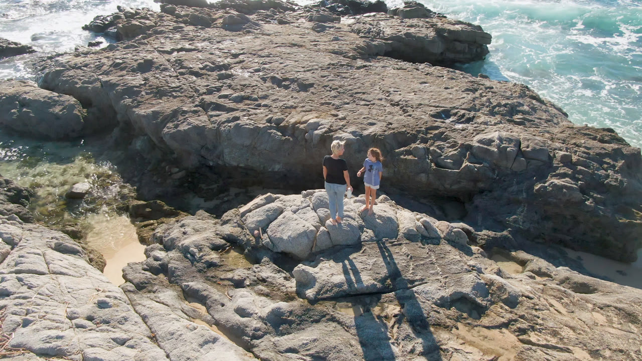 Adult and child stand together on pale rocks along the San Jose Del Cabo coastline, with turquoise water and white surf visible beyond the tide pools.
