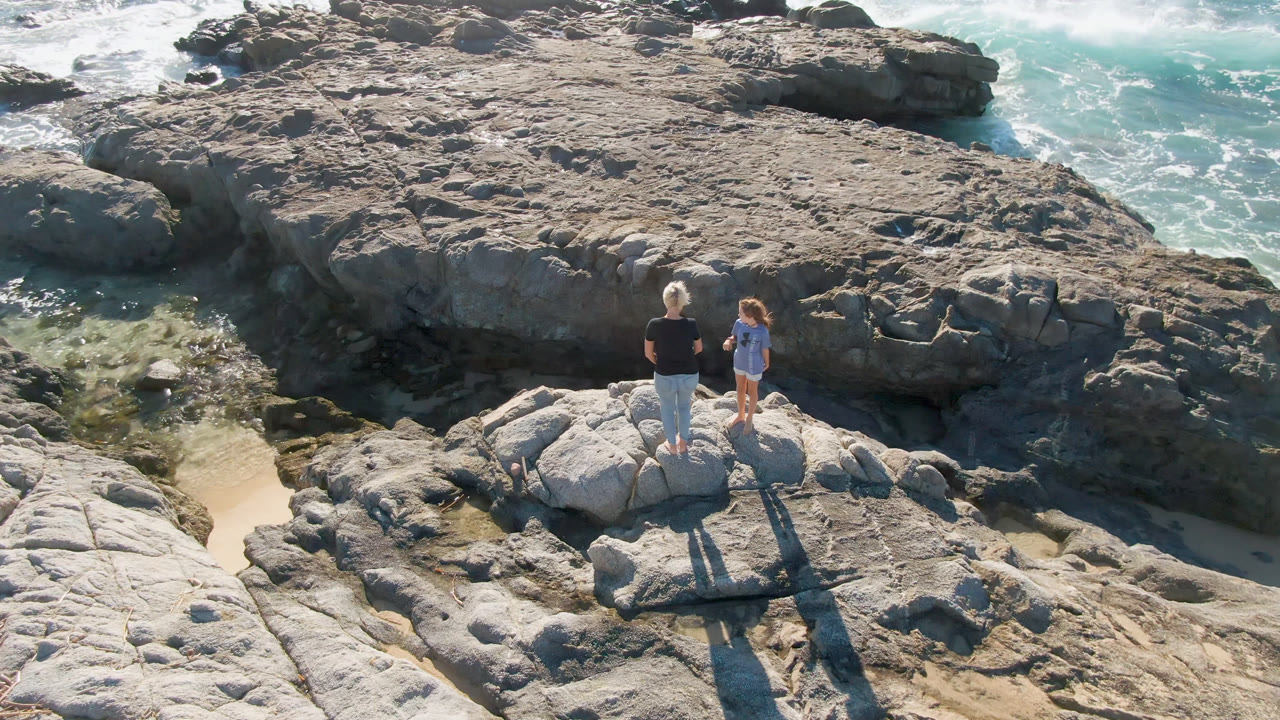 The pair remain on the rocky ledge in San Jose Del Cabo, with the adult facing the child and foamy water moving behind the darker rock shelf.