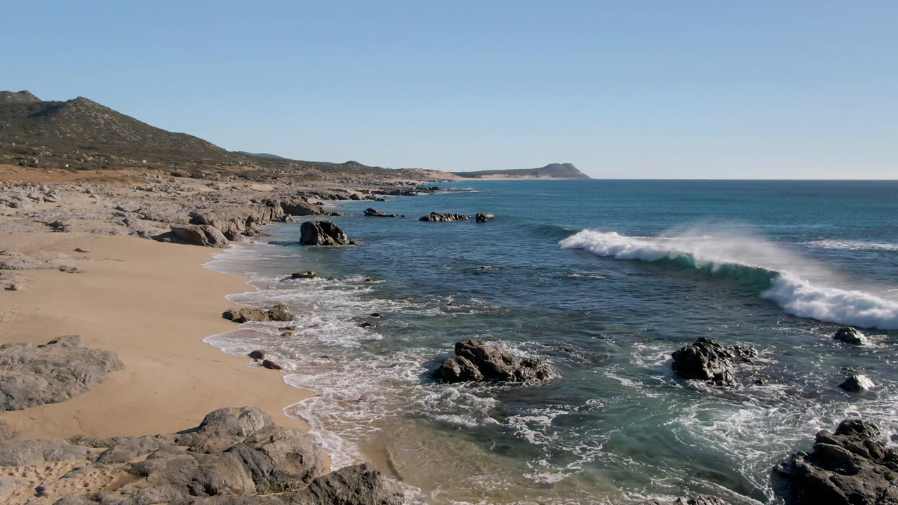 Laguna Beach Los Cabos shoreline with sand at left, dark rocks in shallow water, a breaking wave to the right, low hills behind, and no visible people.